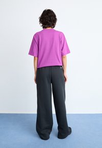 Chemise violette à manches courtes ample, associée à un pantalon noir à jambes larges. L'arrière-plan est minimaliste avec un tapis bleu clair.