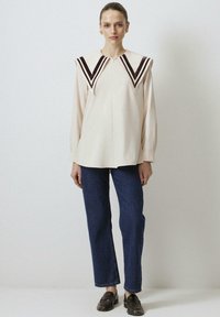Crème lange mouw blouse met een diepe V-vormige kraag, voorzien van donkerbruine fluwelen strepen, gecombineerd met blauwe rechte jeans en zwarte loafers.