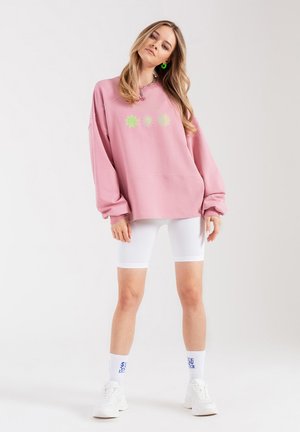 Różowy oversize'owy sweter z trzema zielono-żółtymi wzorami kwiatów, sparowany z białymi rowerowymi szortami z prążkowanej tkaniny i białymi butami sportowymi.