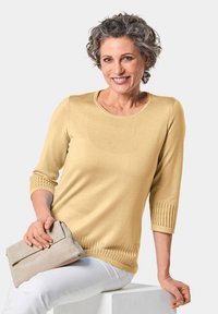 Geel gebreid vest met driekwart mouwen, geribbelde accenten bij de manchetten en zoom, gecombineerd met een lichtgrijze clutch.