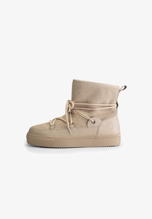 Beige suède enkellaars met dikke rubberen zool, vetersluiting aan de voorkant, een handvat op de hiel en reliëflogo aan de zijkant.