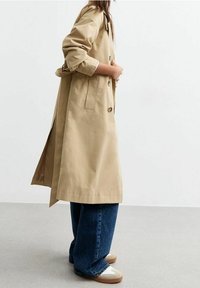 Mujer vistiendo un abrigo trench largo beige, jeans de pierna ancha azules y zapatos color crema, de pie contra un fondo claro y liso.