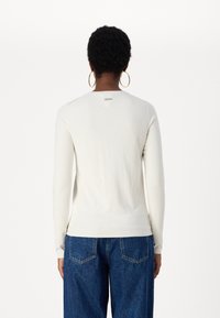 Långärmad, krämfärgad tröja i mjukt material. Har en rundad halsringning och en liten etikett på baksidan. Parat med blåjeans.