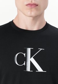 Zwarte katoenen t-shirt met een groot wit en zilverkleurig grafisch logo "cK" en de tekst "Calvin Klein". Ronde halslijn en korte mouwen.