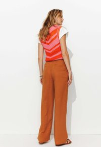Gilet au crochet dans un motif zigzag rose et orange superposé à une chemise blanche, associé à un pantalon large marron, mettant en valeur un tissu texturé.