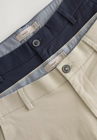Zwei Hosenpaare, eine in Marineblau und eine in Beige, die eine glatte Textur mit Knopfdetails und gemusterten Taillenbünden zeigen. Etiketten sind sichtbar.
