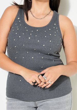 Femme portant un haut gris sans manches orné de petits clous brillants, un collier chaîne argenté, plusieurs bagues en argent et un jean gris clair.