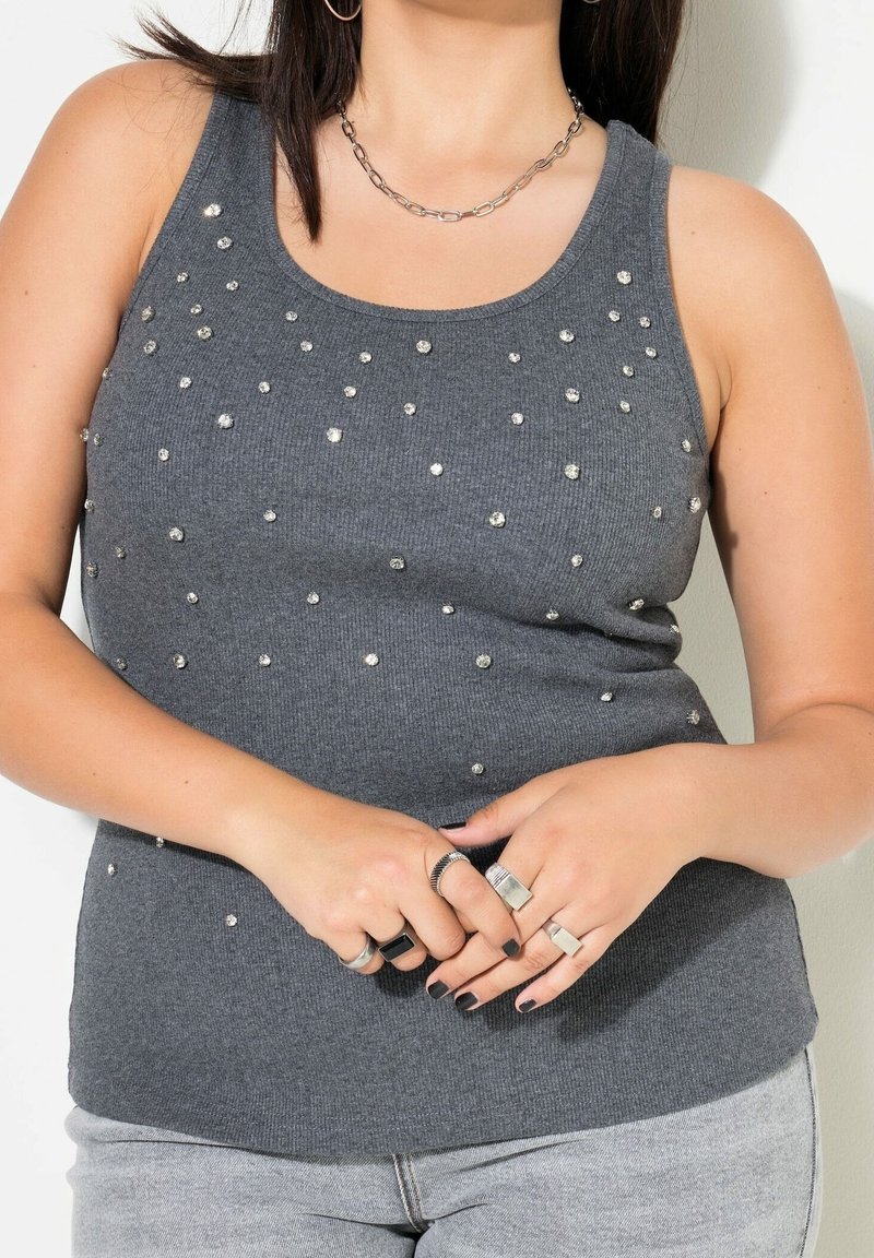Femme portant un haut gris sans manches orné de petits clous brillants, un collier chaîne argenté, plusieurs bagues en argent et un jean gris clair.