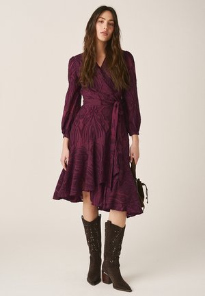 Mujer con un vestido cruzado de manga 3/4 de patrón púrpura y botas negras con tachuelas hasta media pantorrilla, de pie contra un fondo beige.