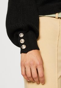 Pull en tricot noir avec une texture côtelée, présentant des boutons argentés sur le poignet. Main visible sur un tissu beige clair.