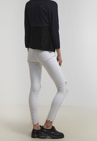 Marinblå kort topp i texturerad svart tyg, i kombination med vita tighta jeans med slitna detaljer och fransade fållar, bärs med svarta skor.