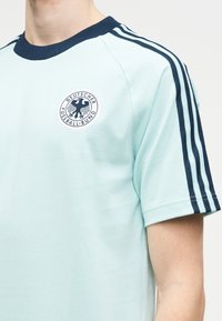 Svetlo modra nemška nogometna majica s temno modrimi ovratnikom, črtami na rokavih in logotipom orla Deutscher Fussball-Bund na levi strani prsnega koša.