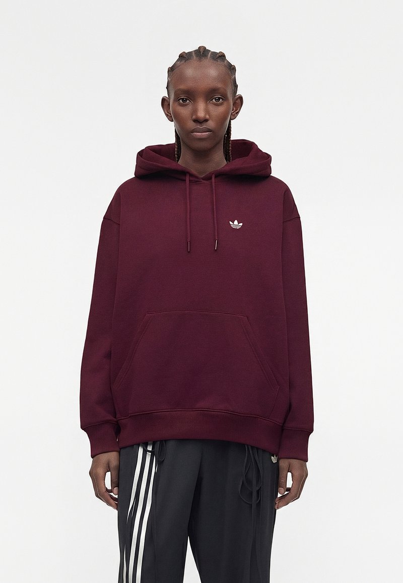 Personne aux cheveux tressés portant un sweat à capuche bordeaux avec une poche avant et un pantalon noir avec des bandes blanches sur les côtés sur un fond uni.