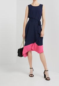 Marinblå klänning med asymmetrisk rosa fåll, ärmlös design och midjebindning. Modellen håller en svart handväska med guldkedja.