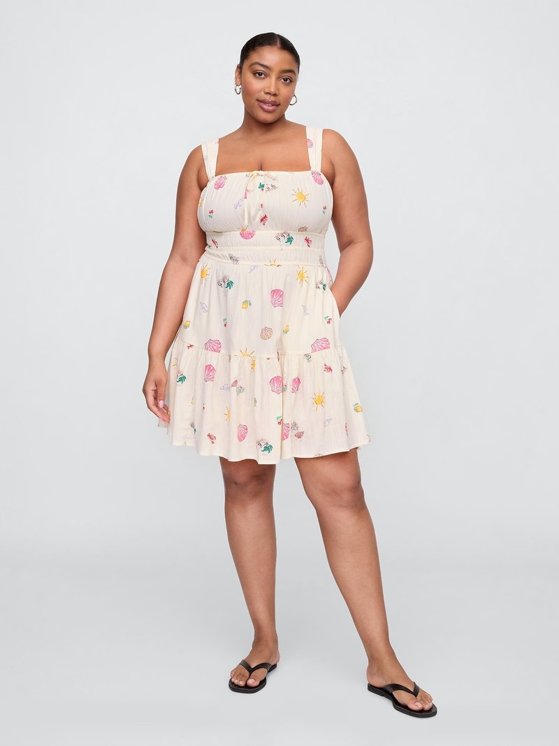 Vrouw in een lichtgekleurde zomerjurk met bloemen- en zonpatronen, met handen in haar zakken, dragend zwarte slippers op een effen achtergrond.