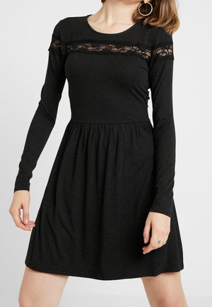 Frau trägt ein schwarzes, langärmliges Kleid mit Spitzenapplikationen über Brust und Schultern, steht vor einem schlichten hellen Hintergrund.