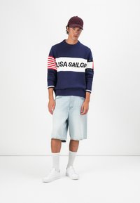 Young man wearing a navy sweater with "USA SAILGP" text, light denim shorts, white sneakers, white socks, and a burgundy cap standing.