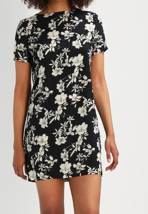 Czarna, krótka sukienka mini z krótkim rękawem i białym kwiatowym nadrukiem, noszona przez osobę stojącą na tle jednolitego jasnego tła.