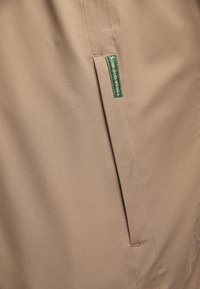 Hnědá tkanina s hladkou texturou, s boční kapsou a zelenou cedulkou s nápisem "Schmuddelwedda." Jednoduchý design a čisté linie.