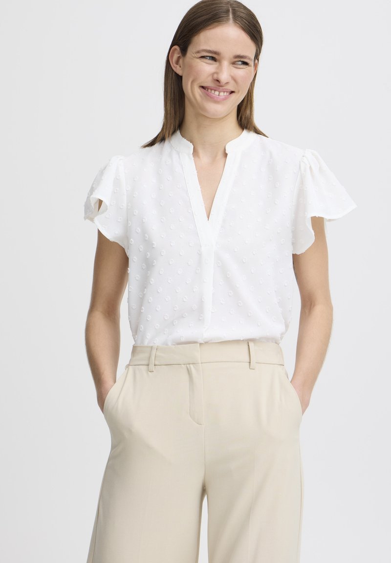 Blusa bianca a maniche corte con un sottile motivo a puntini testurizzato, scollatura a V e maniche arricciate, abbinata a pantaloni beige su misura.