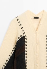 Light beige button-up garment with black and brown fabric panels and black stitched detailing along seams and edges.