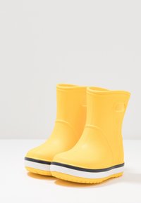 Bottes en caoutchouc jaunes à la cheville avec une texture lisse, un bout rond, une languette de tirage au talon et une semelle rayée blanche et noire.