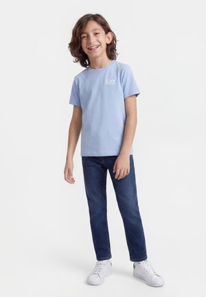 Garçon souriant aux cheveux mi-longs portant un t-shirt EA7 bleu clair, un jean bleu foncé et des baskets blanches, debout devant un fond blanc.