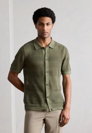 Άνδρας που φοράει πουκάμισο πλεκτό με κοντό μανίκι σε ελιά πράσινο χρώμα με μοτίβο δίχτυ και μπεζ παντελόνι, στέκεται μπροστά σε απλό φωτεινό φόντο.