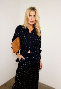 Ξανθιά γυναίκα που φοράει μαύρο πουκάμισο με κουμπιά και παντελόνι με μικρές κίτρινες πουά, κρατώντας κάτω από το χέρι της ένα καφέ τσαντάκι.