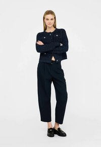 Tamsiai mėlynas megztas kardiganas su auksinio tono sagomis, derinamas su laisvo silueto tamsiomis kelnėmis ir juodais loaferiais, pabrėžiant tekstūruotą audinį.