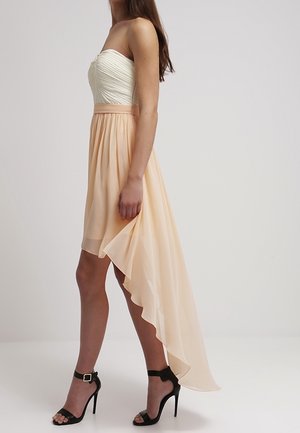 Frau trägt ein schulterfreies, weiß-rosa asymmetrisches Kleid mit fließendem, hoch-tief geschnittenem Saum und schwarzen, hochhackigen Sandalen mit Knöchelriemen.