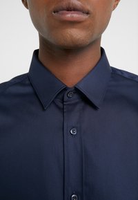 Gros plan d'une personne portant une chemise boutonnée bleu foncé avec un col pointu, ne montrant que le bas du visage et le cou.