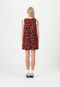 Vestido sin mangas de tela negra con un patrón de corazones rojos y pequeños puntos. Presenta un corte holgado y un dobladillo redondeado. Se lleva con calcetines blancos y zapatos negros.
