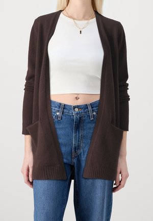 Cardigan lungo marrone realizzato in un tessuto morbido, con scollatura aperta, polsini a costine e due tasche frontali. Indossato sopra un crop top bianco e jeans.