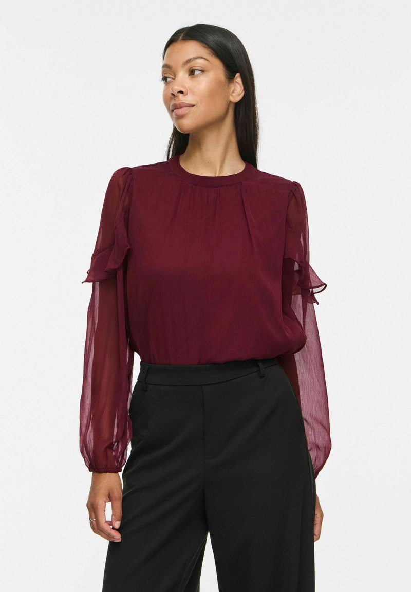 Femme portant un chemisier transparent bordeaux à manches longues avec des volants, associée à un pantalon noir taille haute, regardant vers la gauche sur un fond uni.