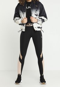 Giacca bomber bianca e nera con design astratto, leggings neri con aperture e sneakers nere con dettagli dorati.