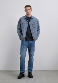 Joven de pie con las manos en los bolsillos, vistiendo una chaqueta de mezclilla azul clara, camiseta negra, vaqueros azules y zapatos negros, contra un fondo liso.