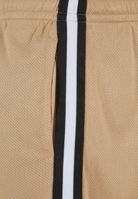 Shorts en mesh beige avec une surface texturée, présentant une bande noire et blanche sur le côté. Comprend un détail de poche latérale.