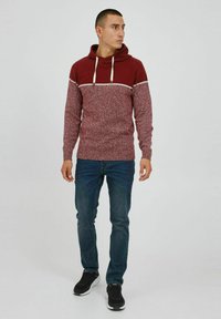 Haut à capuche en tricot bordeaux avec une finition texturée, bande horizontale blanche sur la poitrine, associé à un jean bleu et des baskets noires.