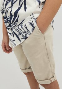 Camicia a maniche corte con un motivo a foglie bianco e navy, abbinata a pantaloni corti beige arrotolati con una tasca laterale e dettagli di cucitura discreti.