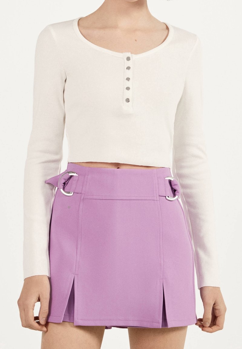 Femme portant un haut court à manches longues blanc avec des boutons et une jupe taille haute violette avec des détails de boucles sur les côtés et des fentes à l'avant.