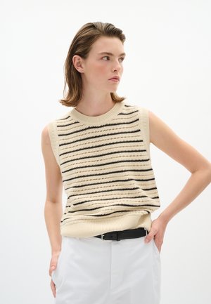 Person trägt einen ärmellosen cremefarbenen Stricktop mit schwarzen Streifen und weiße Hose, steht mit einer Hand in der Tasche und schaut zur Seite.