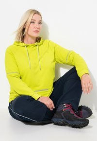 Felpa verde lime con cordini e tasca frontale; abbinata a pantaloni navy con dettagli bianchi. Stivali da escursione neri con lacci rossi.