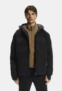 Melns pūkains mētelis ar kapuci, ar izšūtu tekstūru, kas aprīkots ar rāvējslēdzēju un regulējamām aprocēm, vilkts virs brūna augsta kakla topa.