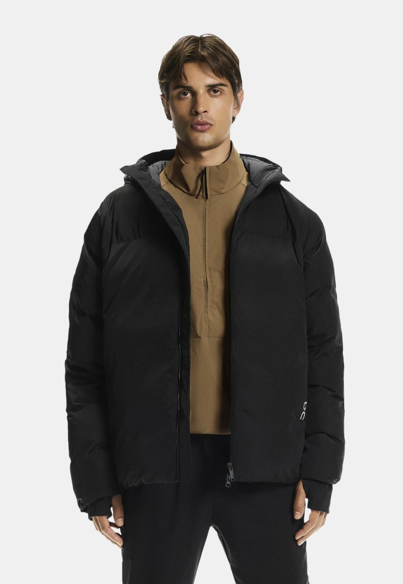 Schwarze Pufferjacke mit Kapuze, gesteppte Textur, mit Reißverschluss und verstellbaren Bündchen, über einem braunen Shirt mit hohem Kragen getragen.