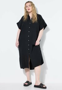 Vestido negro, de corte amplio, con botones en la parte frontal y mangas cortas, y aberturas laterales. La tela lisa y los botones blancos destacan el diseño minimalista.