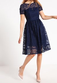 Abito in pizzo blu navy con maniche corte, motivo floreale, corpetto aderente e gonna con orlo a taglio. Abbinato a tacchi rosa chiaro.