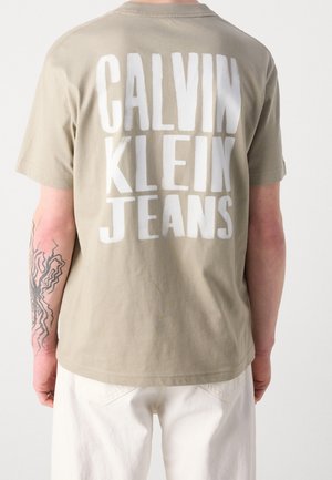 Persoon draagt een beige T-shirt met korte mouwen met grote witte tekst "CALVIN KLEIN JEANS" op de achterkant, gecombineerd met witte broek en een zwarte tatoeage op de arm zichtbaar.