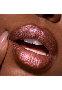 Metallic pink læbestift på fulde læber, med en glat tekstur og blank finish. Hvide tænder og en neutral baggrund fremhæver detaljerne.