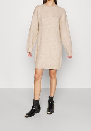 Person trägt ein beiges, langärmeliges Strickkleid und schwarze Knöchelstiefel mit metallischen Spitzen, steht vor einem einfarbigen Hintergrund.